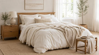 Cozy bedroom with layered cream bedding, textured throw blanket, wooden nightstand, and potted plant.